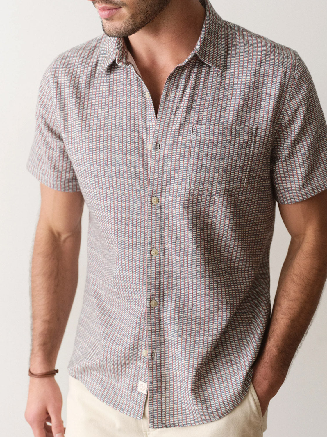 A man models the Marine Layer Stretch Selvage Short Sleeve Shirt, featuring a classic fit and subtle checkered pattern, with one hand in his pocket against a plain background. His face is partially out of frame.