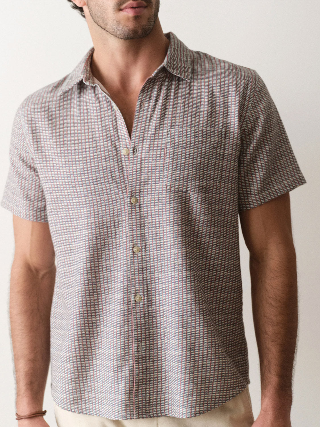A man stands against a plain background wearing the Marine Layer Stretch Selvage Short Sleeve Shirt, featuring a classic collar, relaxed fit, and subtle muted plaid pattern; his face is partially out of frame.