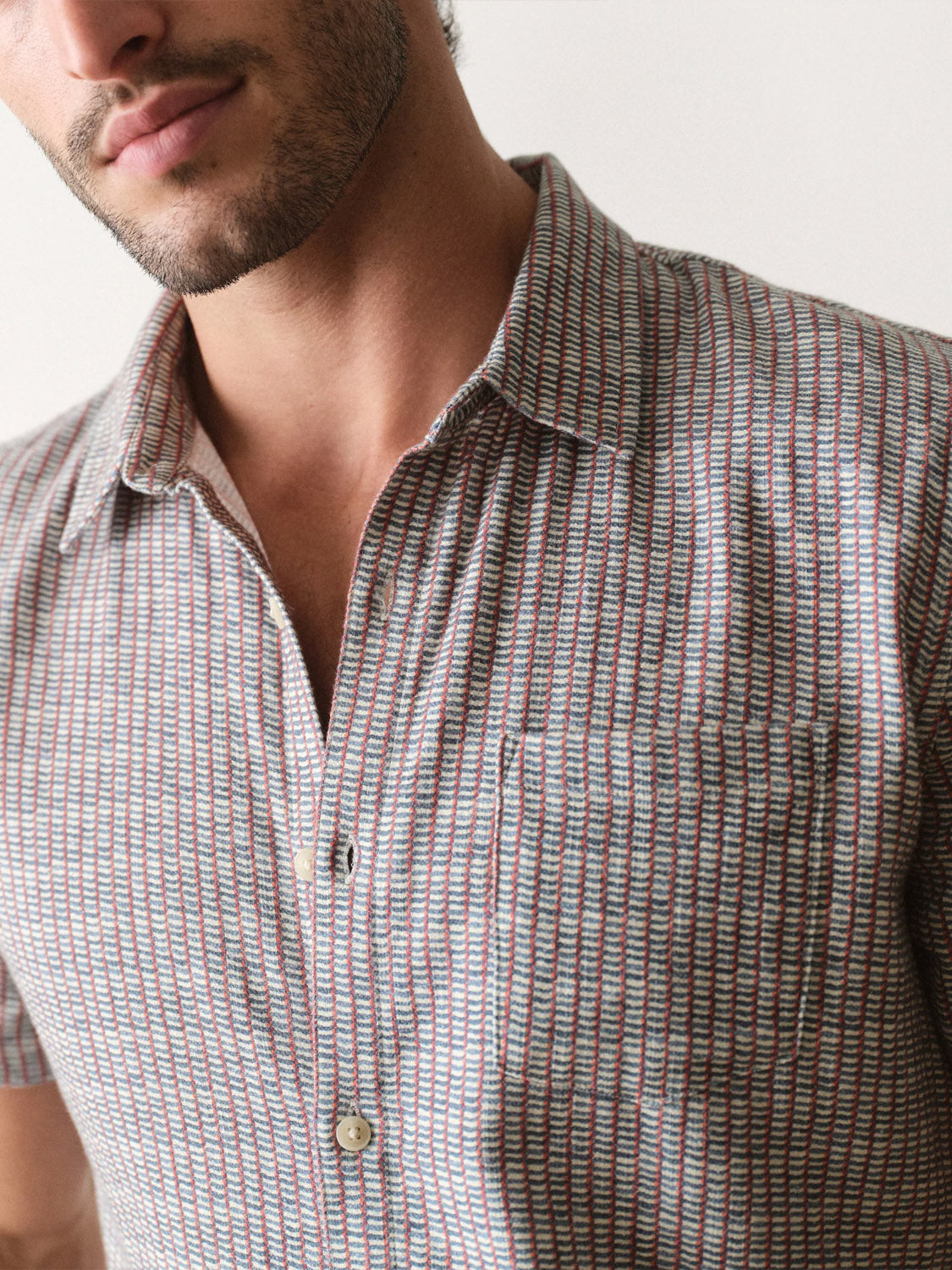 A close-up of a man wearing the Marine Layer Stretch Selvage Short Sleeve Shirt, featuring a classic fit, chest pocket, and small red, blue, and white geometric pattern. Two cream-colored buttons are visible; his face is partially shown.