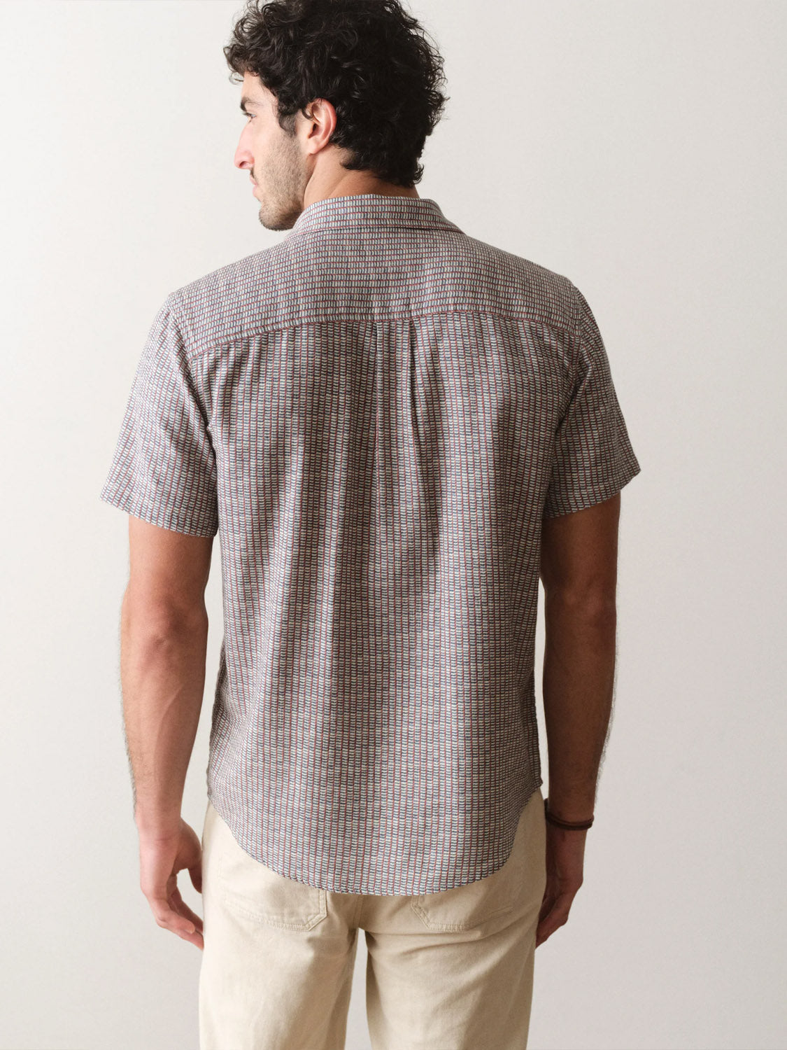A man with curly dark hair is seen from behind, wearing the Marine Layer Stretch Selvage Short Sleeve Shirt and light-colored pants, standing against a plain light background.