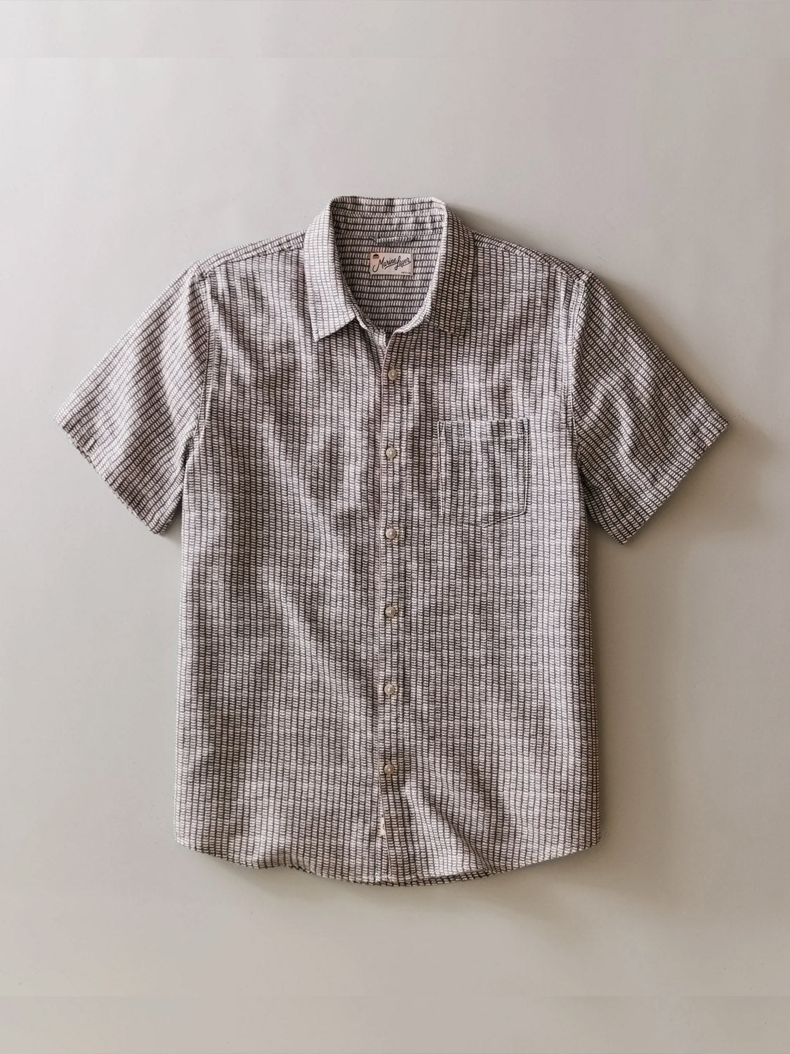 The Marine Layer Stretch Selvage Short Sleeve Shirt, featuring a classic fit and left chest pocket, is shown laid flat. It has a stretchy button-down design with a subtle gray and white checkered pattern.