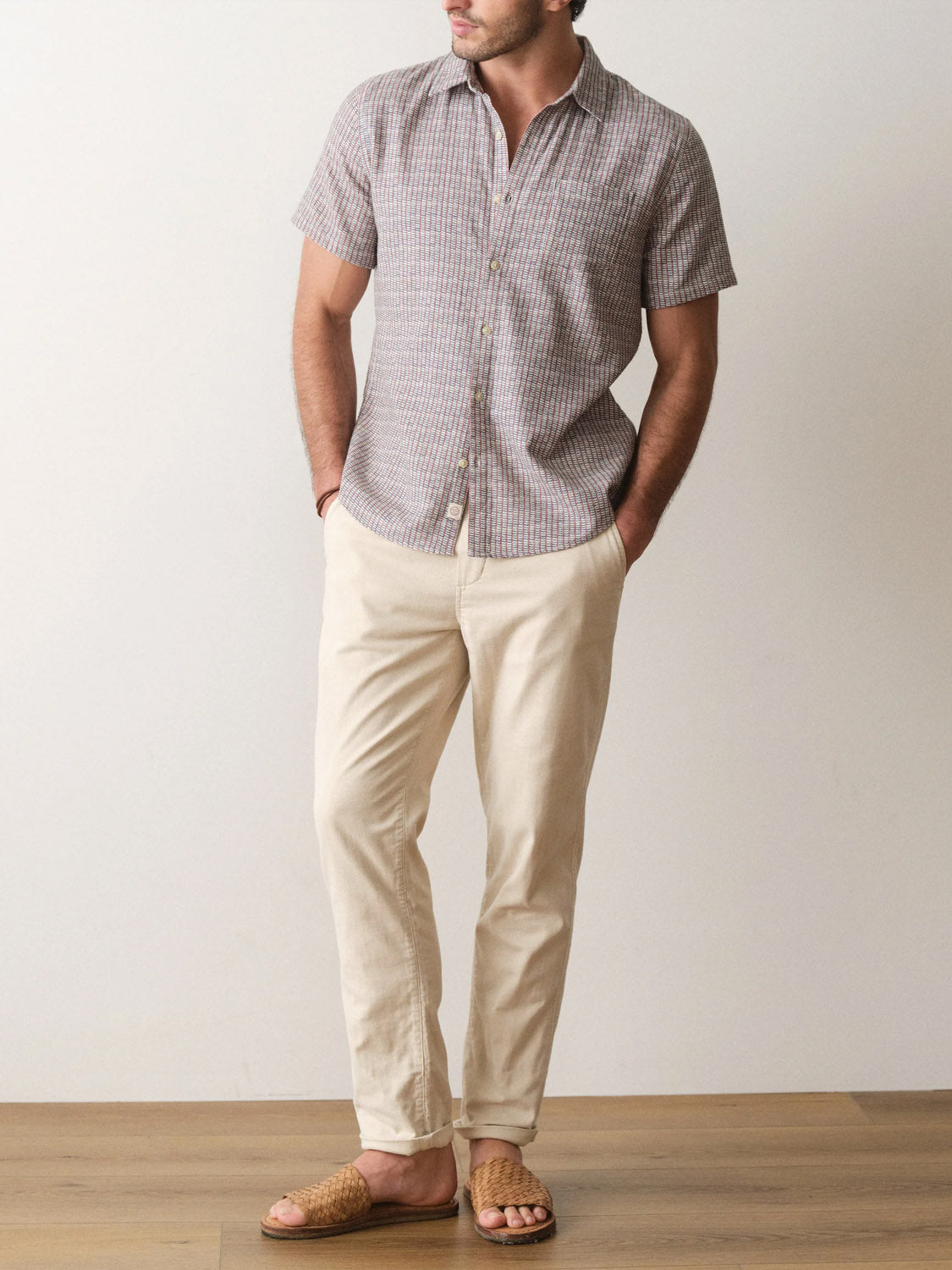 A man stands indoors with his hands in his pockets, wearing the Marine Layer Stretch Selvage Short Sleeve Shirt, light beige pants, and woven brown sandals.