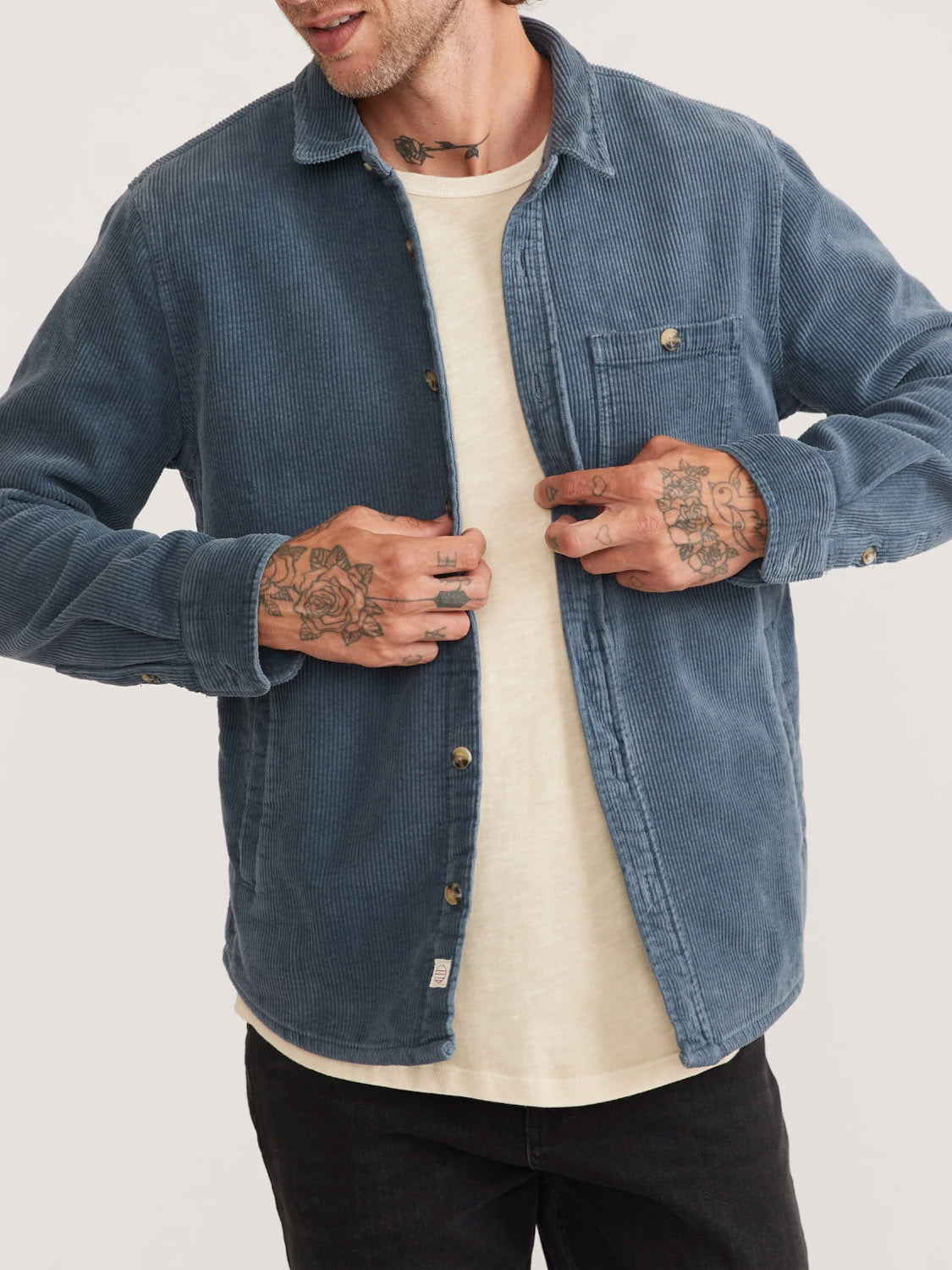 A man adjusts his open Marine Layer Max Broken In Corduroy Overshirt in blue, revealing a white T-shirt underneath. His hands and wrists are covered in tattoos, and hes wearing dark pants. His face remains out of view.