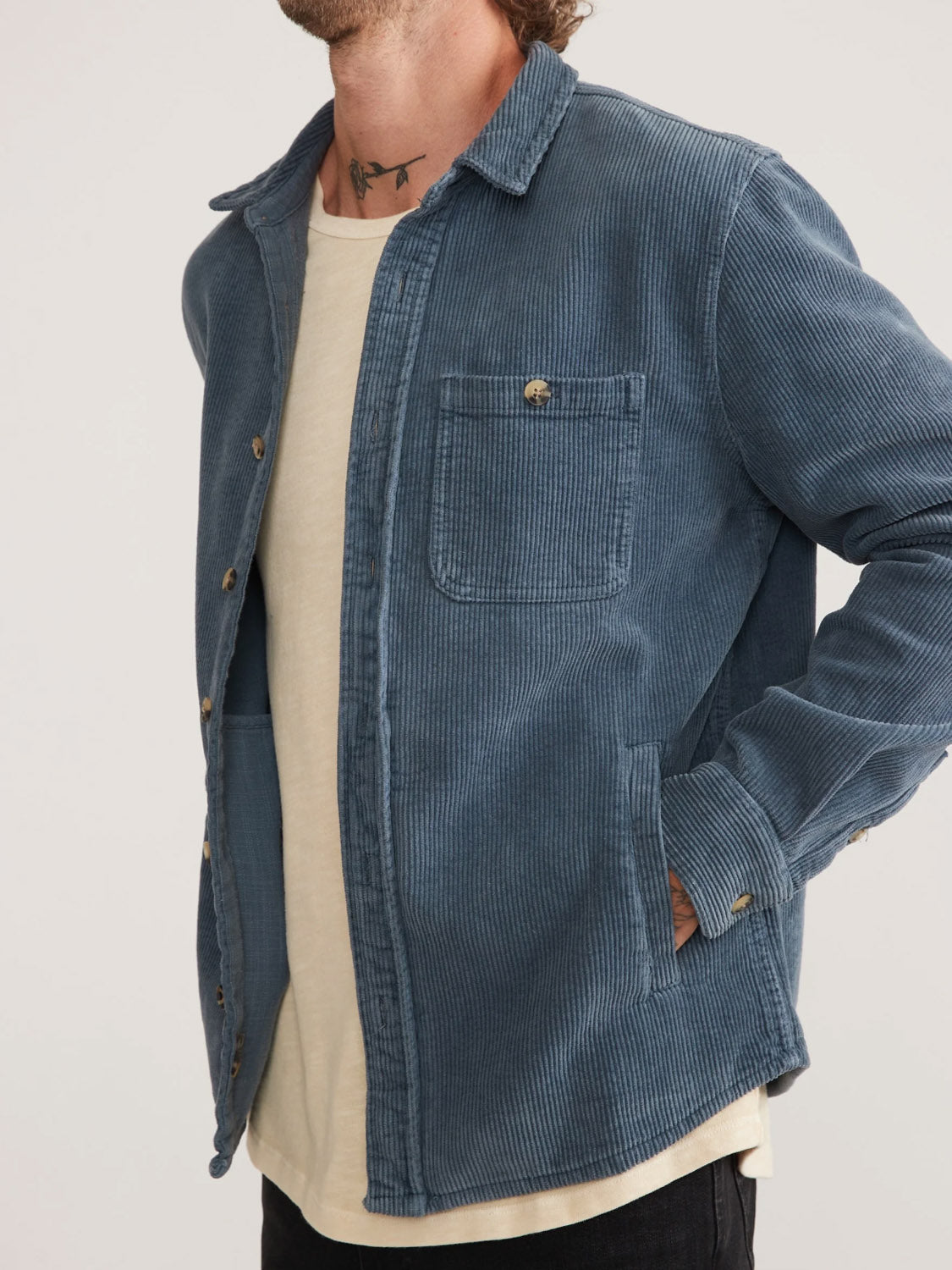 Someone is wearing a blue Marine Layer Max Broken In Corduroy Overshirt over a beige T-shirt, with hands in pockets. The overshirt has a single chest pocket and buttoned front. A neck tattoo is visible, with the image focusing on the torso and not showing the face.