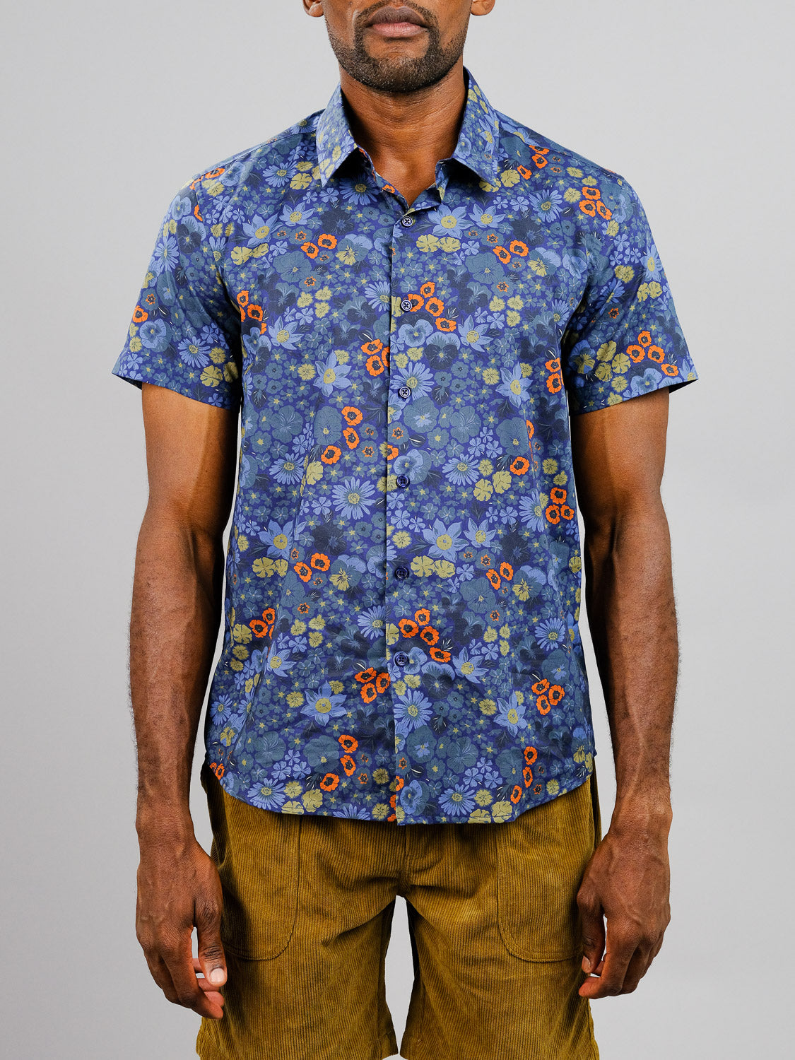 A man wears the Meadow Printed Cotton Shirt by ID Menswear—featuring a vibrant floral pattern and short sleeves—paired with mustard pants, standing against a plain light gray background.