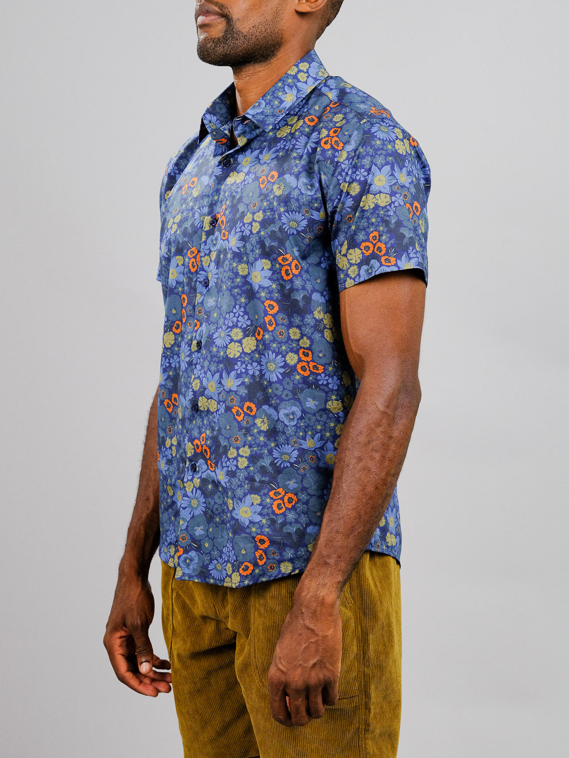 A man wears the Meadow Printed Cotton Shirt by ID Menswear and mustard yellow corduroy pants, standing slightly to the side against a plain gray background.