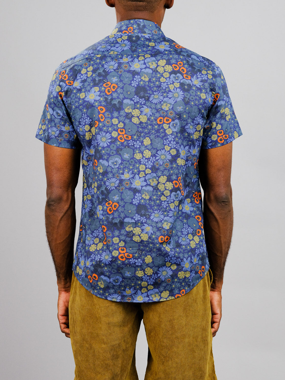 A person stands with their back to the camera, wearing the Meadow Printed Cotton Shirt by ID Menswear—featuring vibrant blue floral prints with red and yellow accents—paired with mustard yellow corduroy pants against a light gray background.