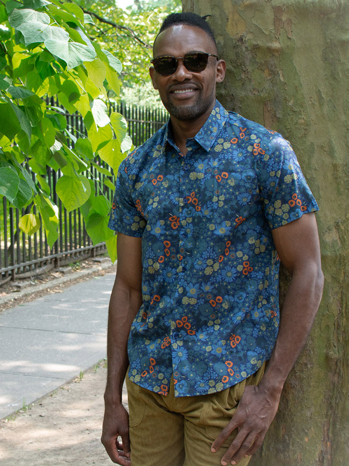 A man smiles by a leafy tree and black metal fence on a sunny day, wearing sunglasses and the Meadow Printed Cotton Shirt from ID Menswear—a lightweight blue floral cotton shirt.