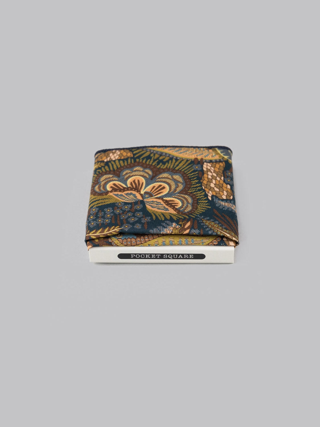 The Liberty™ Medusa Pocket Square by ID Menswear, handmade in Brooklyn from fine Tana Lawn cotton with blue, green, and gold patterns, is folded atop a box labeled POCKET SQUARE against a plain gray background.