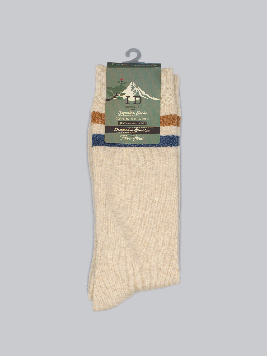 A pair of warm, beige Rutland Cotton Melange Socks by ID Menswear with blue and brown stripes at the top hang on a hanger against a light gray background. The packaging label features a mountain logo and reads Superior Socks, Cotton Merino, marking them as winter essentials for premium comfort.