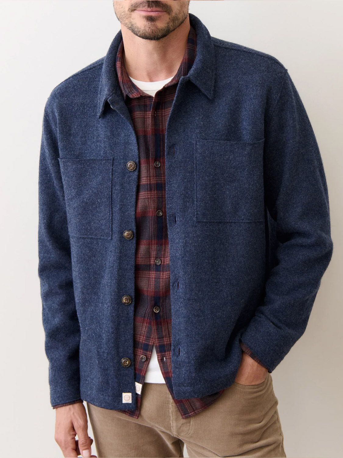 A man stands against a plain light-colored wall, wearing Marine Layer's Mendocino Wool Overshirt in blue over a red and black plaid shirt with tan pants. His left hand is in his pocket and his face is partially visible.