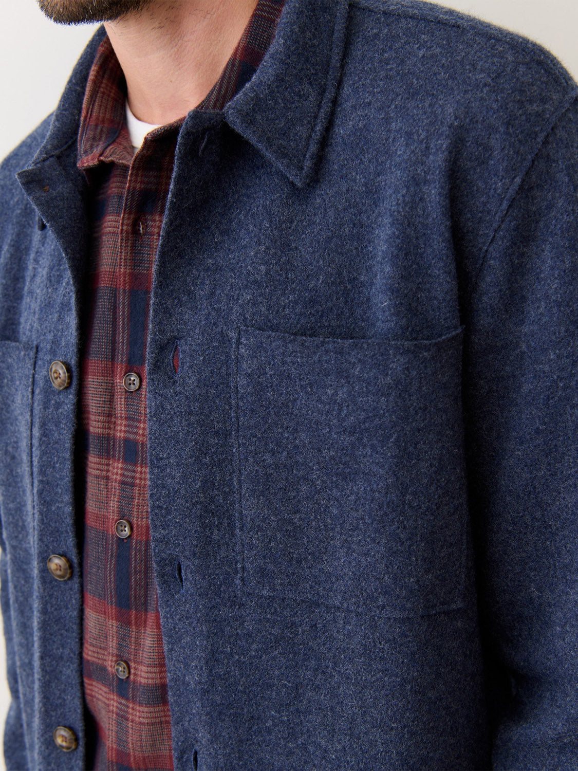 A close-up shows a person wearing the Marine Layer Mendocino Wool Overshirt in navy blue over a red and navy plaid shirt, highlighting the chest, face, and details like the overshirt’s buttons and pockets.