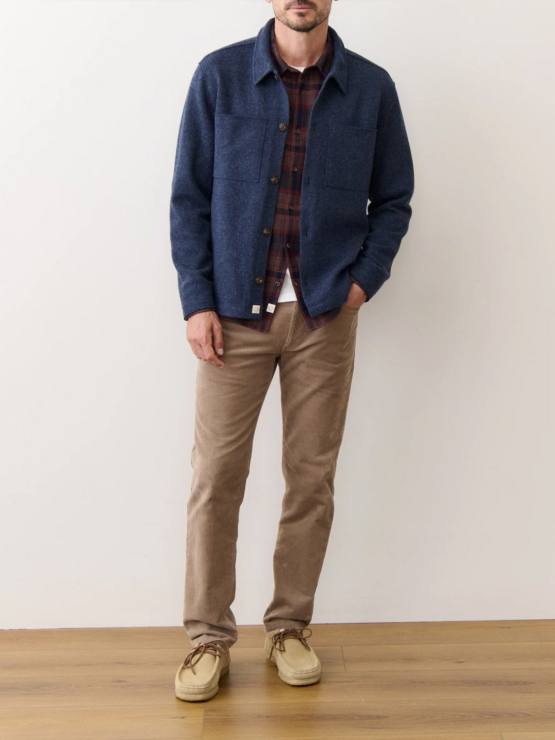A man stands indoors wearing the Marine Layer Mendocino Wool Overshirt over a red plaid shirt, with beige pants and tan suede shoes. His left hand is in his pocket, face partially out of frame, against a plain white wall and wood floor.