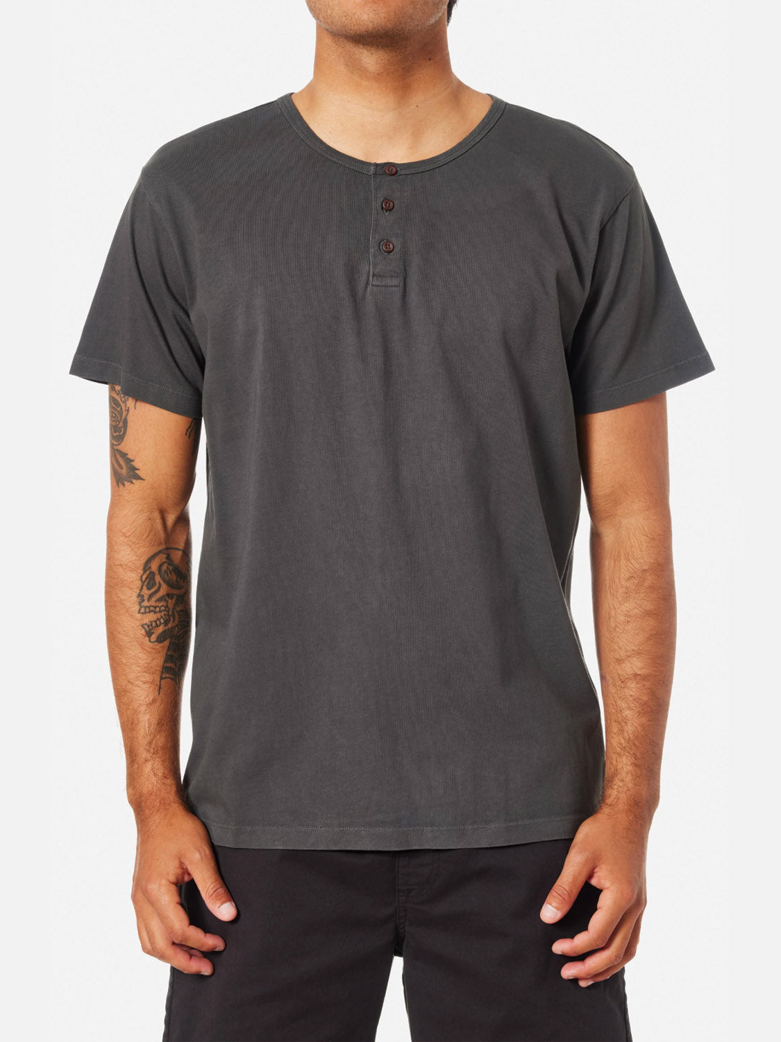 A man stands with relaxed arms, wearing the Katin Mesa Henley—a short-sleeved, dark grey men’s shirt with three buttons at the collar—showing tattoos on his left arm and paired with black pants.