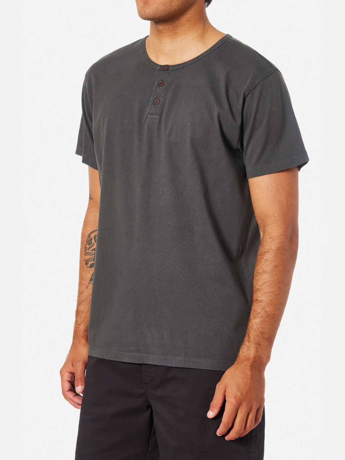 A man wearing the Katin Mesa Henley in dark gray with three buttons, paired with black shorts. He has a tattoo on his left forearm and stands against a plain white background.