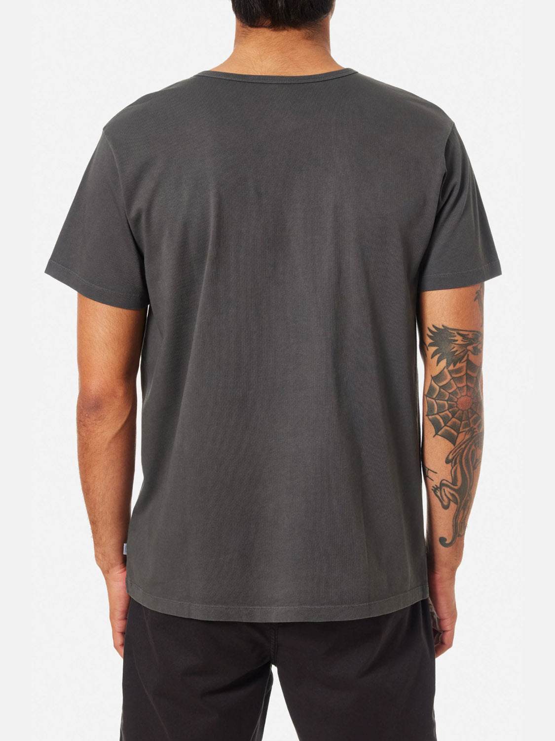 A person seen from behind wears a Katin Mesa Henley in dark gray and black shorts, showing a large dragon tattoo on their left forearm and upper arm.