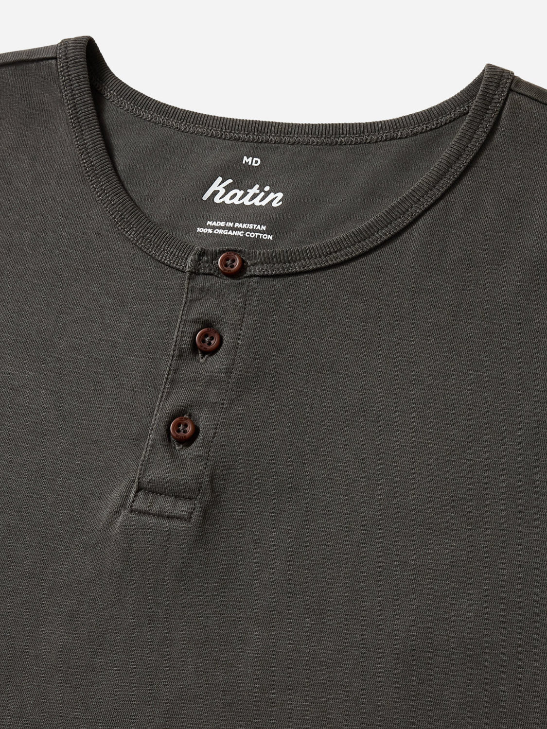 A close-up of Katin's Mesa Henley in dark gray highlights the collar, three brown buttons, and a white men’s medium label. Made in Mexico from 100% organic cotton, it’s ideal as your everyday henley.