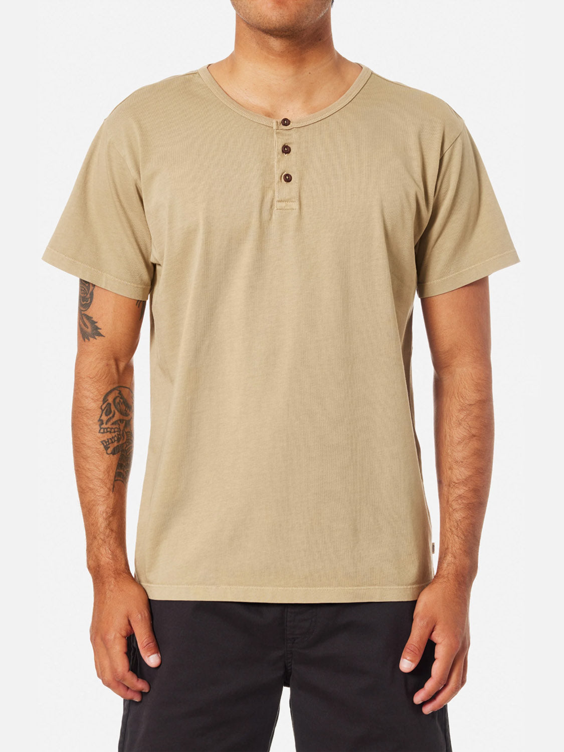 A man wears the Katin Mesa Henley, an organic cotton shirt with a three-button collar, and black shorts. His tattooed forearms are visible as he stands against a plain white background, showcasing this casual everyday look.