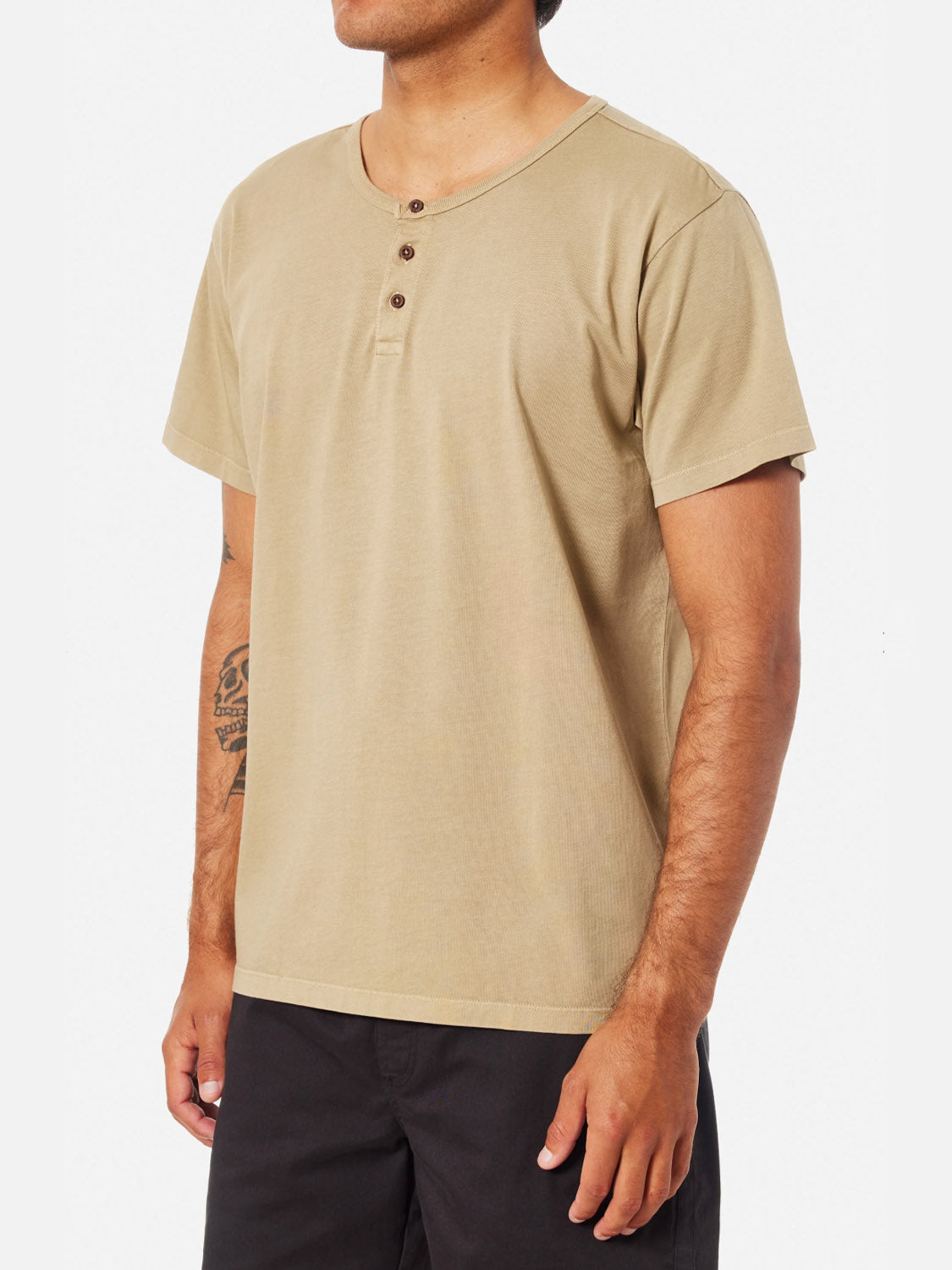 A man wears the Katin Mesa Henley in light brown with three buttons at the collar and black pants, standing against a plain white background. A tattoo on his left forearm adds personality to the look.