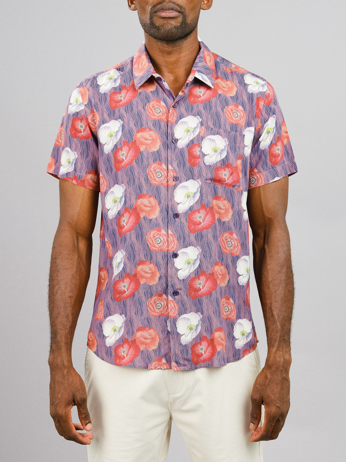 A man in the Metok Printed Rayon Shirt by ID Menswear, featuring vibrant red, white, and purple florals, paired with cream pants, stands against a plain light gray background.