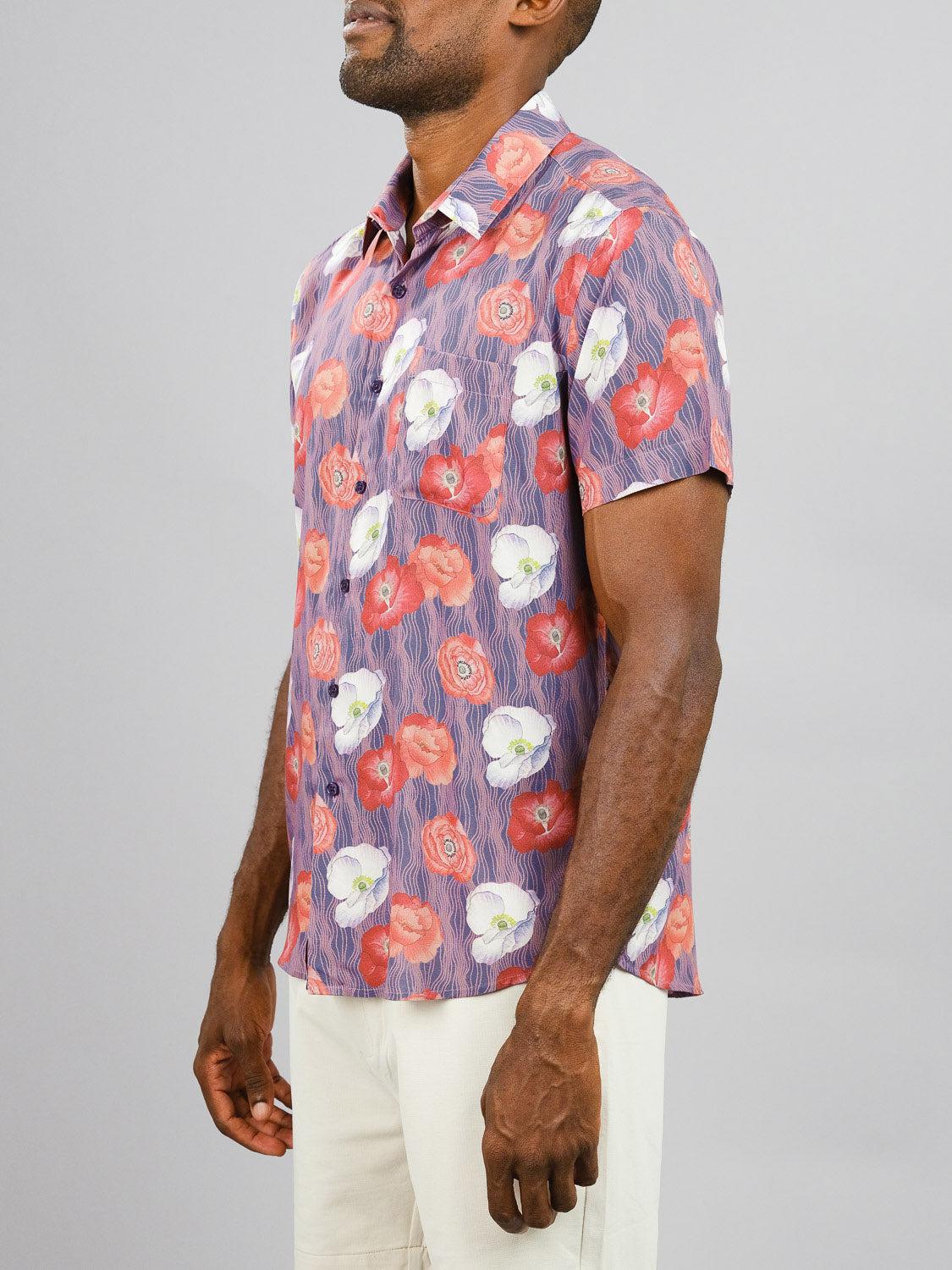 A man wears the Metok Printed Rayon Shirt by ID Menswear, featuring a vibrant red, white, and purple floral pattern, paired with light pants against a plain grey background—ideal for warm weather.