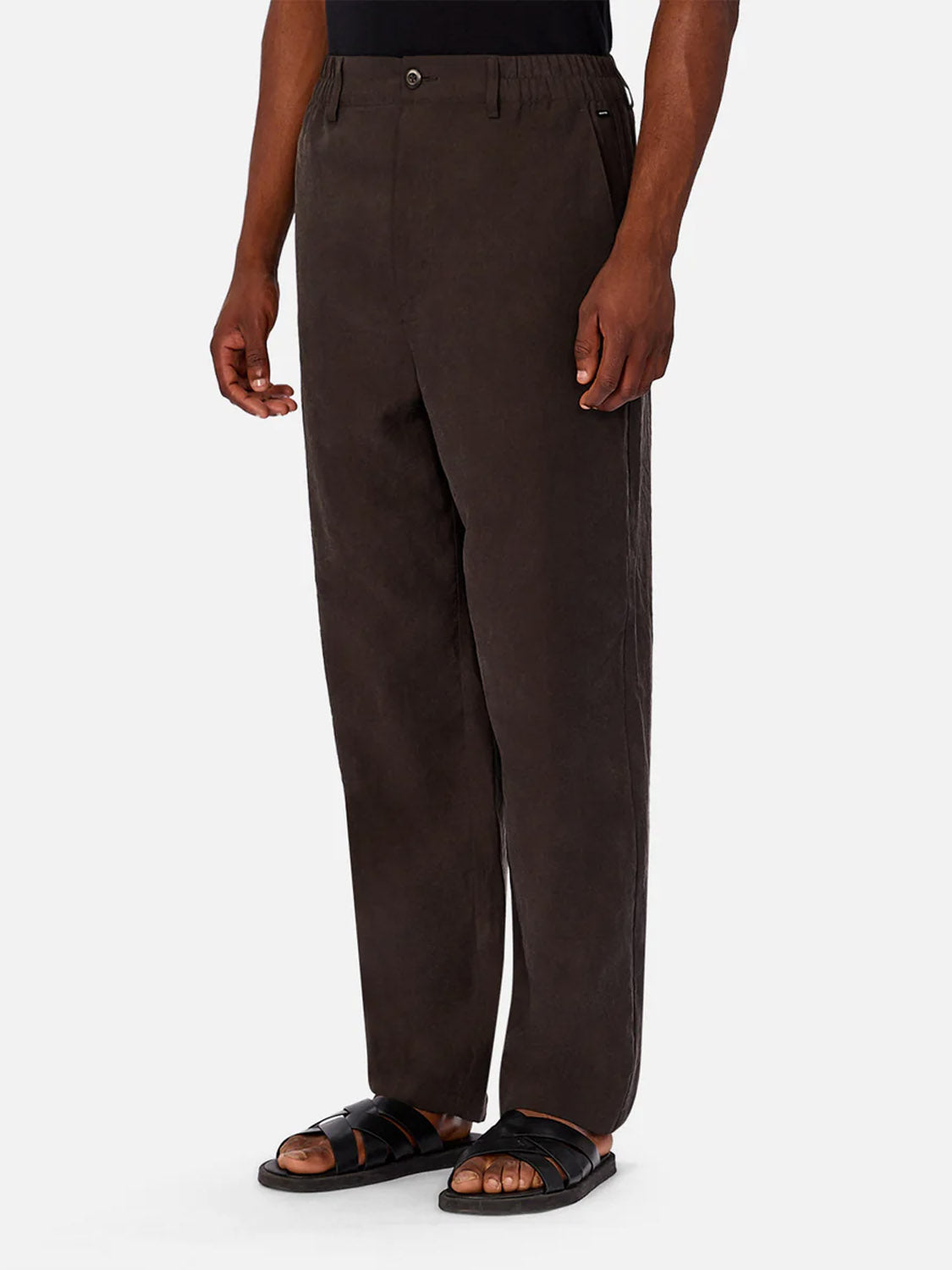 A person wears Industrie’s The Miami Pant in dark brown, paired with a black shirt and black sandals, standing against a plain light gray background with arms relaxed at their sides.
