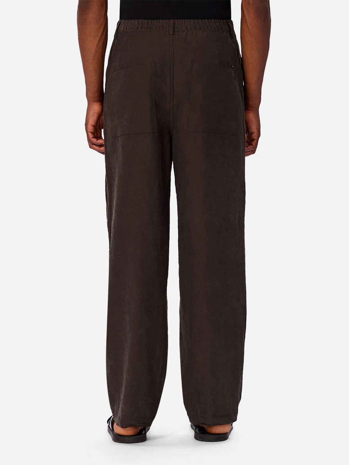 A person faces away, wearing The Miami Pant by Industrie—loose-fitting, dark brown lyocell-blend pants with an elastic waistband and back patch pockets—paired with black sandals.