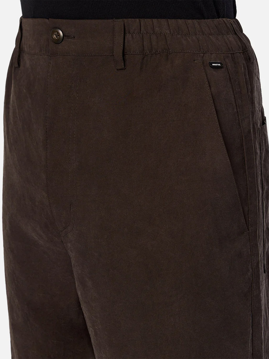 A close-up of a person wearing The Miami Pant by Industrie—dark brown men's straight leg trousers featuring an elastic waistband, side pockets, button closure, and a small black tag by the pocket, paired with a tucked-in black shirt.