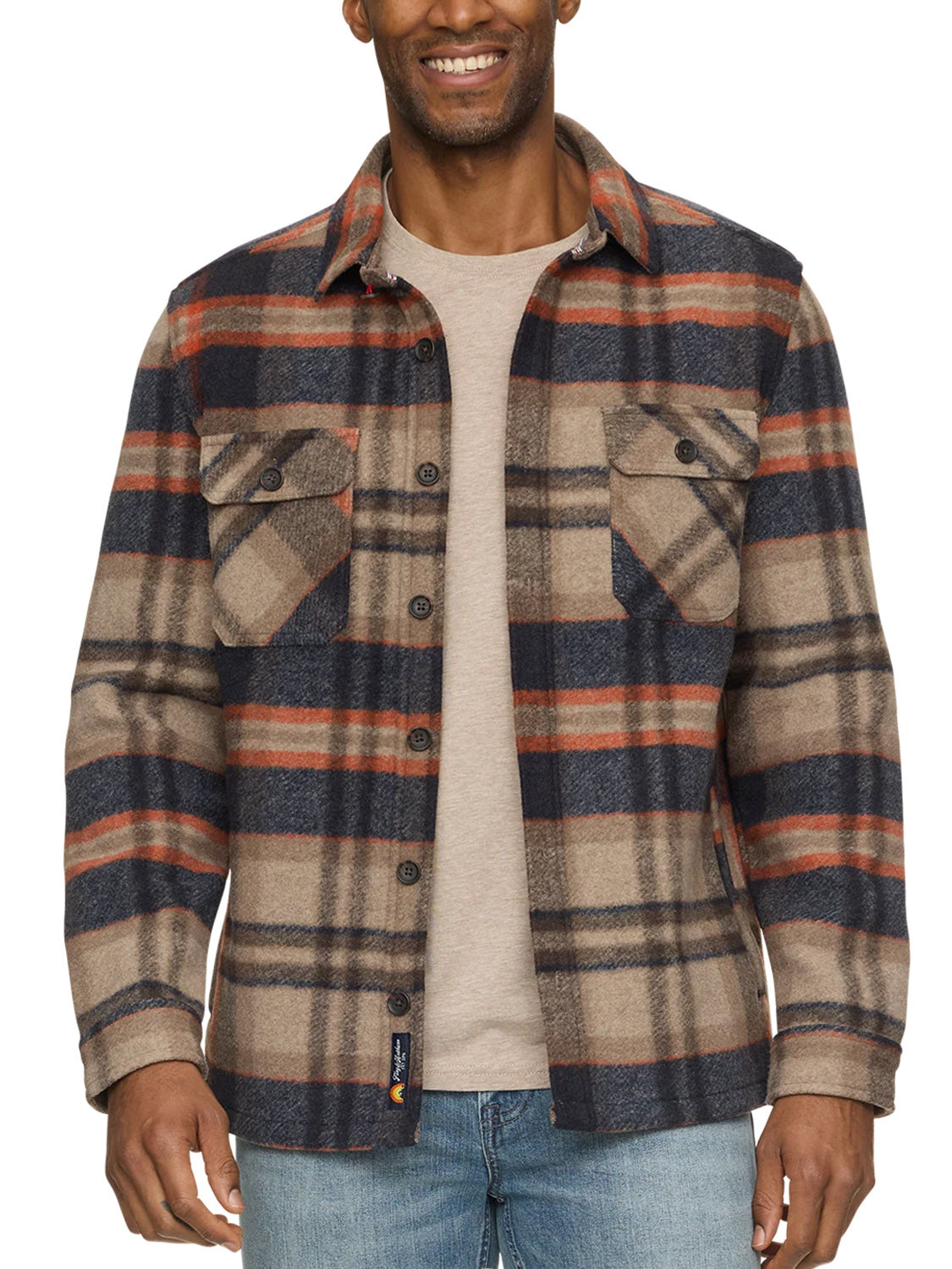 A man wearing the Flag and Anthem Midland Jacket—a beige, navy, and red plaid brushed shirt jacket—over a light tan t-shirt and blue jeans, smiles with his hands relaxed by his sides.