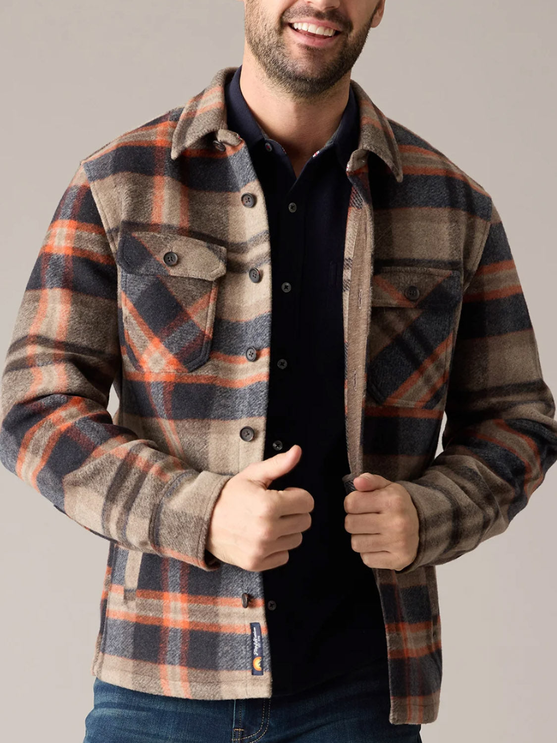 A man in the Flag and Anthem Midland Jacket—plaid in beige, orange, and navy—smiles as he adjusts the brushed fabric over a black shirt, showing this men’s jacket is perfect for casual, comfortable layering.