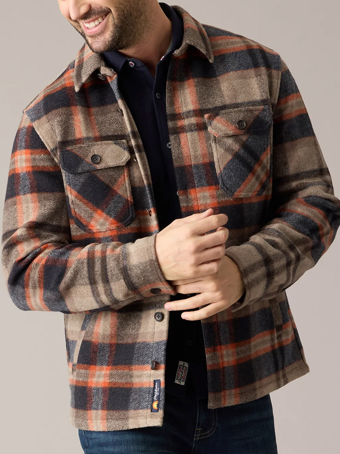 A man smiles as he buttons the Flag and Anthem Midland Jacket, featuring soft, brushed plaid fabric in brown, beige, navy, and orange. His head is slightly turned to the side.