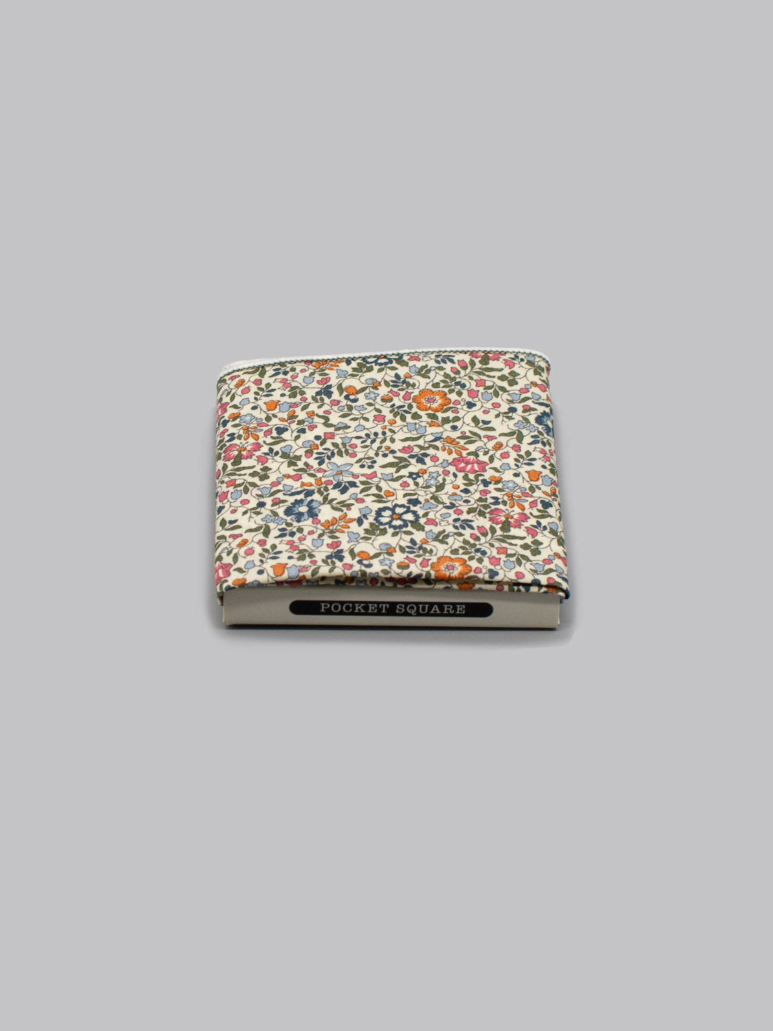 The Liberty™ Mille Pocket Square by ID Menswear, handmade in Brooklyn from Liberty of London Tana Lawn, features a floral pattern with red, orange, and blue flowers and sits partially tucked into a box labeled POCKET SQUARE on a gray background.