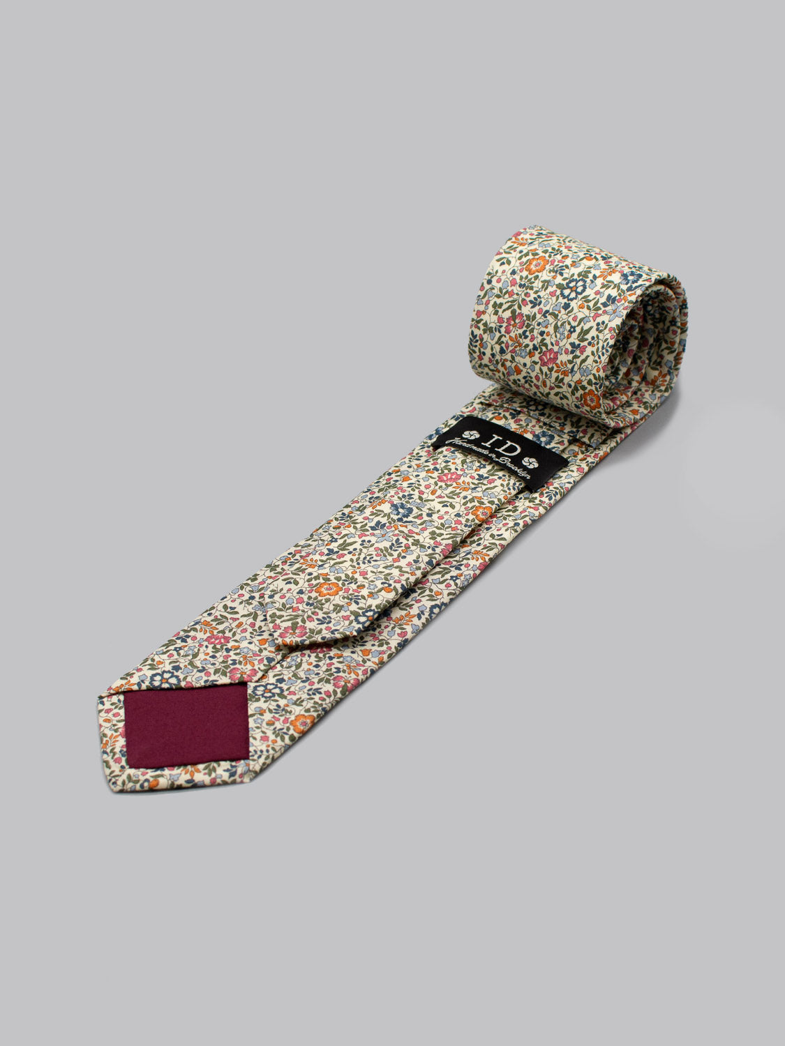 A rolled-up Liberty of London Tana Lawn™ Millie tie by ID Menswear, handmade in Brooklyn from floral cotton with a visible brand label and solid maroon tip, is displayed on a plain gray surface.