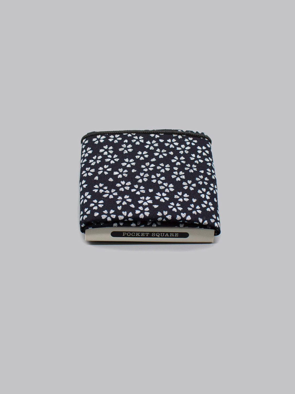 The Mini Bloom Pocket Square by ID Menswear is a handmade black cotton square featuring small white floral patterns, shown folded against a gray background with a label reading “Pocket Square.”.