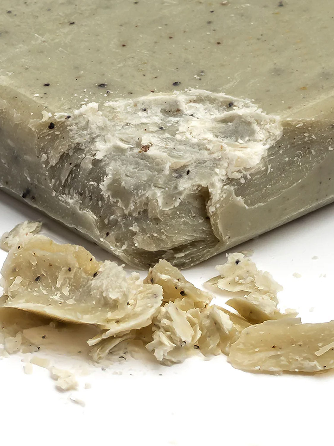 Close-up of a partially used Exfoliating Mini Body Bar by Baxter, beige with small black specks. Nearby, soap shavings lie scattered on a white surface, releasing hints of cedarwood and oakmoss.