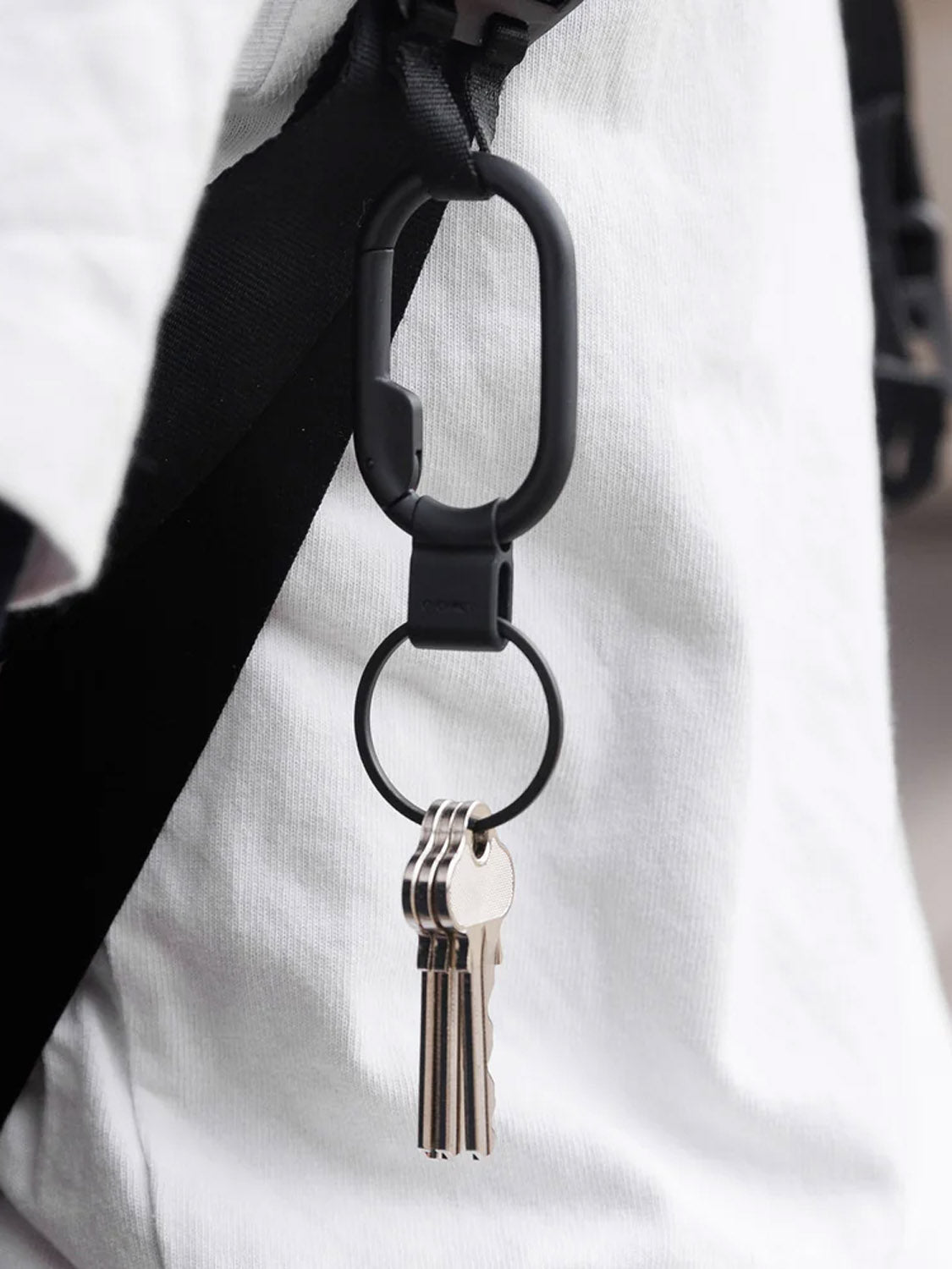 A close-up of the Orbitkeys Clip Mini Keyring, a minimalist carabiner-style keychain, clipped to a black strap and holding three silver keys. Part of a white shirt is visible in the background.