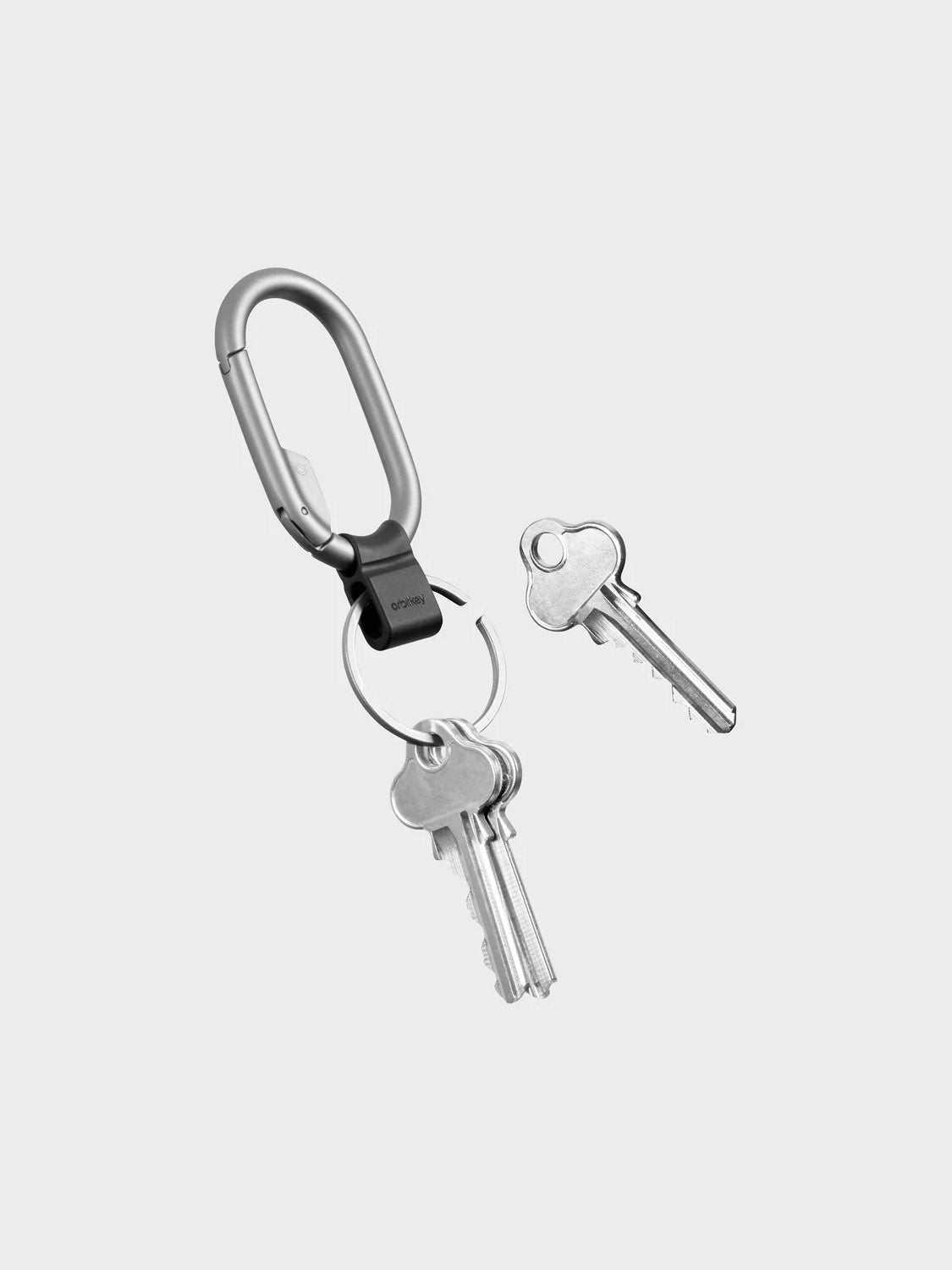 A set of silver keys on the Orbitkeys Clip Mini Keyring featuring a minimalist carabiner clip, plus one loose key nearby, displayed on a plain light gray background.