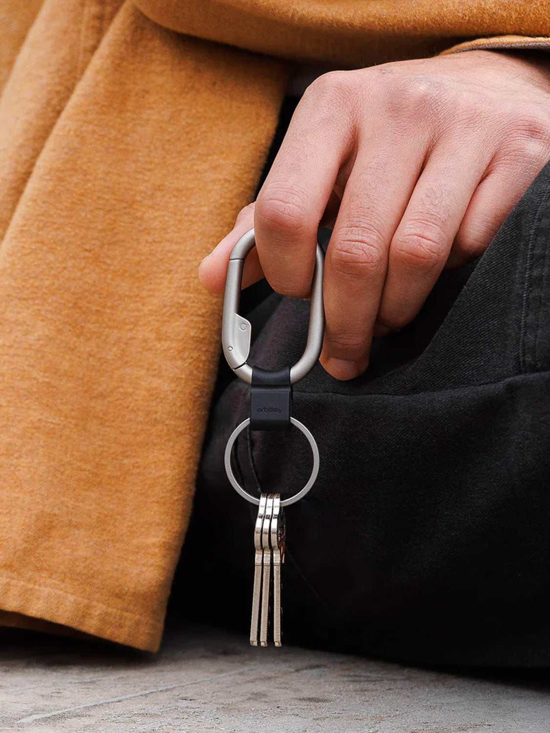 A person in a tan coat and black pants holds the Orbitkeys Clip Mini Keyring with keys attached, their hand resting near their knee.