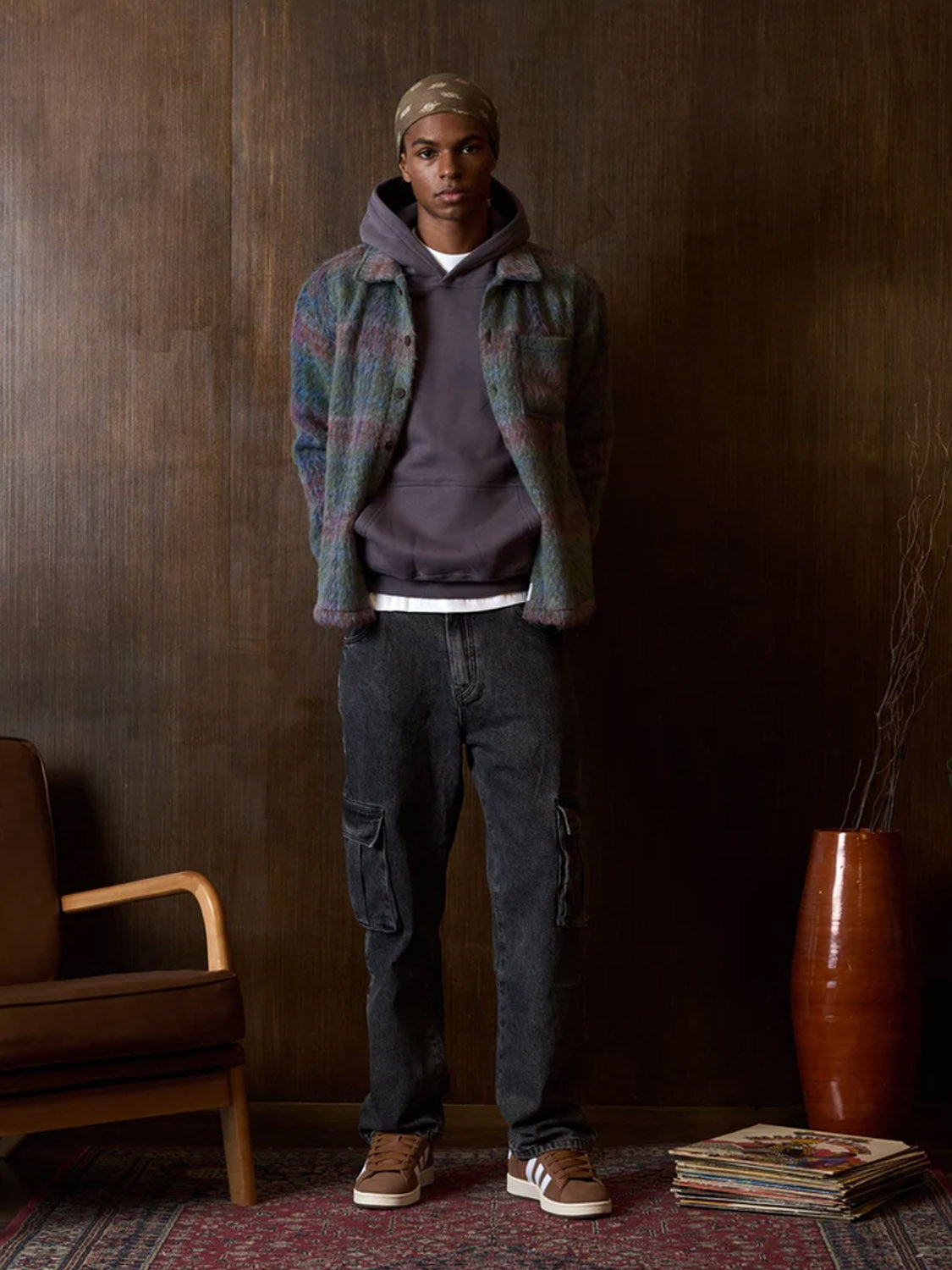 Indoors, a person wears the Kuwalla Faux Mohair Shirt with a classic collar, layered over a dark hoodie, paired with black cargo jeans and brown sneakers. Nearby are a chair, vase, and stack of magazines on a patterned rug.