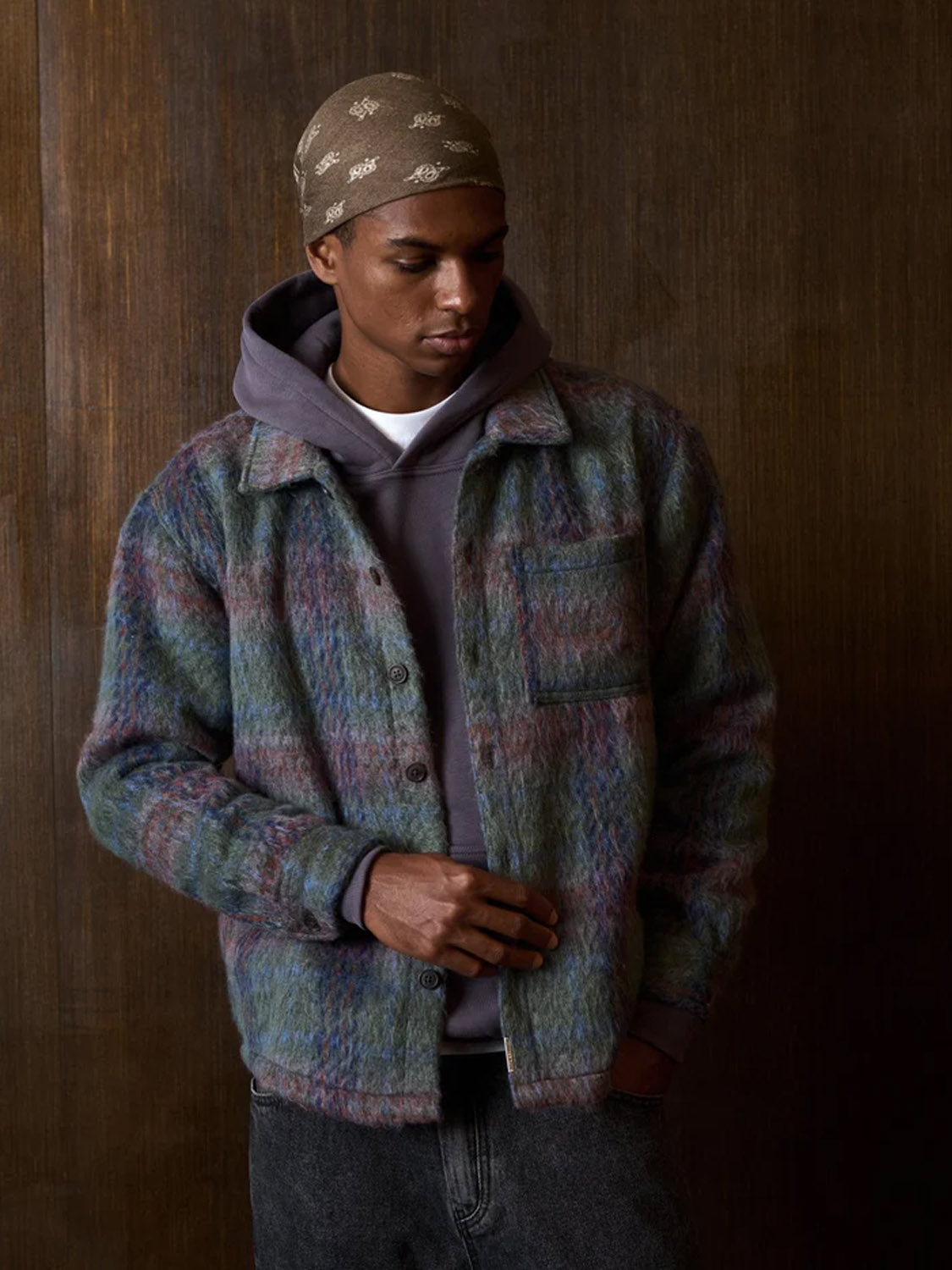 A person wearing a Kuwalla Faux Mohair Shirt with a classic collar, layered under a multicolored jacket and purple hoodie, paired with a patterned beige bandana and jeans, stands against a wooden background looking down.