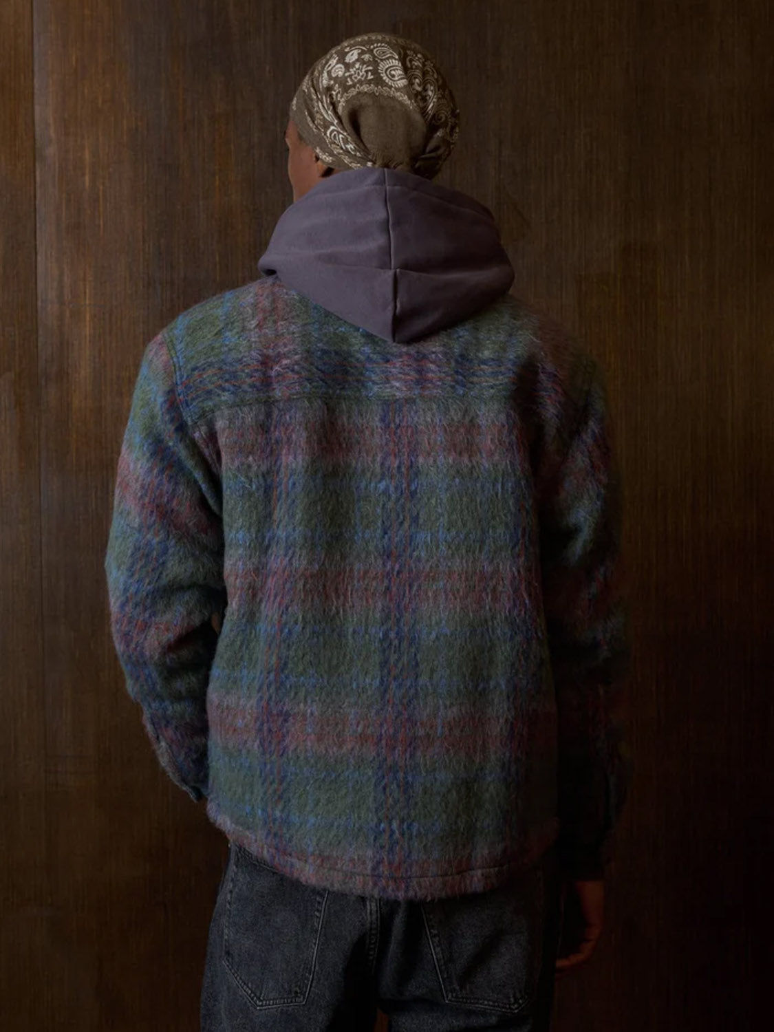 A person faces away from the camera, wearing Kuwalla's Faux Mohair Shirt—a multicolored plaid piece with a classic collar and chest pocket—layered over a gray hoodie, paired with dark jeans and a brown bandana, against a wooden backdrop.