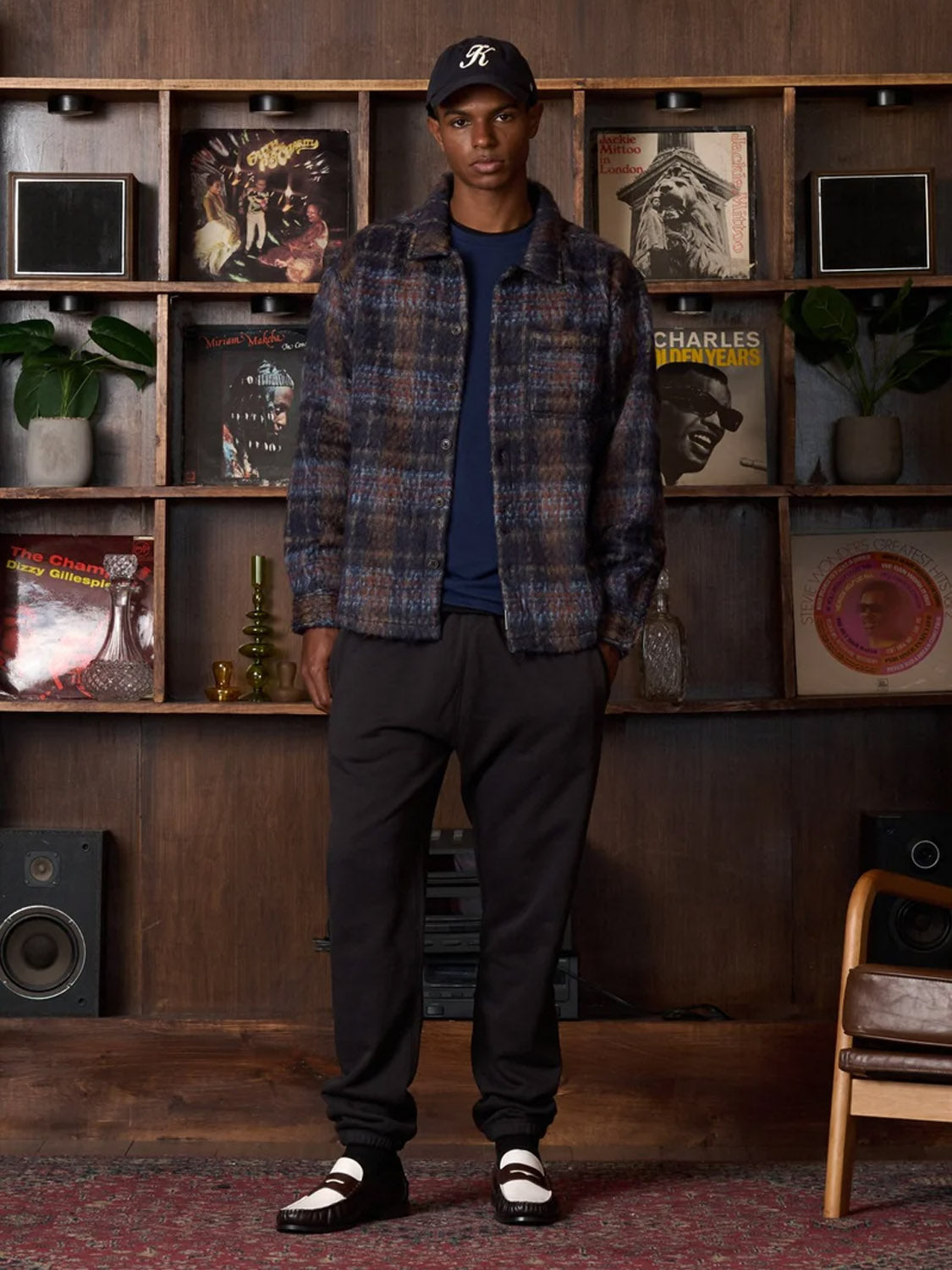 A man stands by a bookshelf filled with records, speakers, and plants, wearing the Kuwalla Faux Mohair Shirt with a classic collar and chest pocket, along with a plaid jacket, black pants, two-tone loafers, and a black baseball cap.
