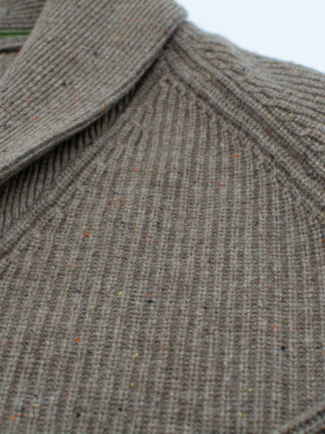 Close-up of a textured, ribbed fabric with a shawl collar in neutral taupe. The Montreal Merino Wool Cardigan from ID Menswear is crafted with speckled yarn, featuring small hints of color for a subtle, dotted pattern that enhances its cozy appeal.