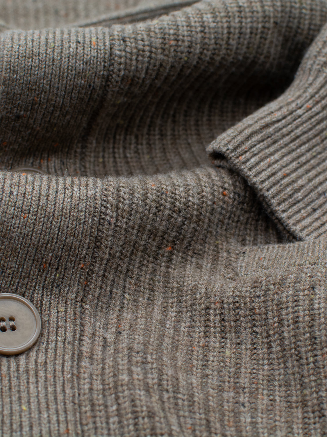 Close-up of the Montreal Merino Wool Cardigan by ID Menswear, showcasing a textured gray knit with vertical ribbing made from speckled yarn. Multicolored flecks enhance its look, while a partially sewn brown button highlights its fine craftsmanship.