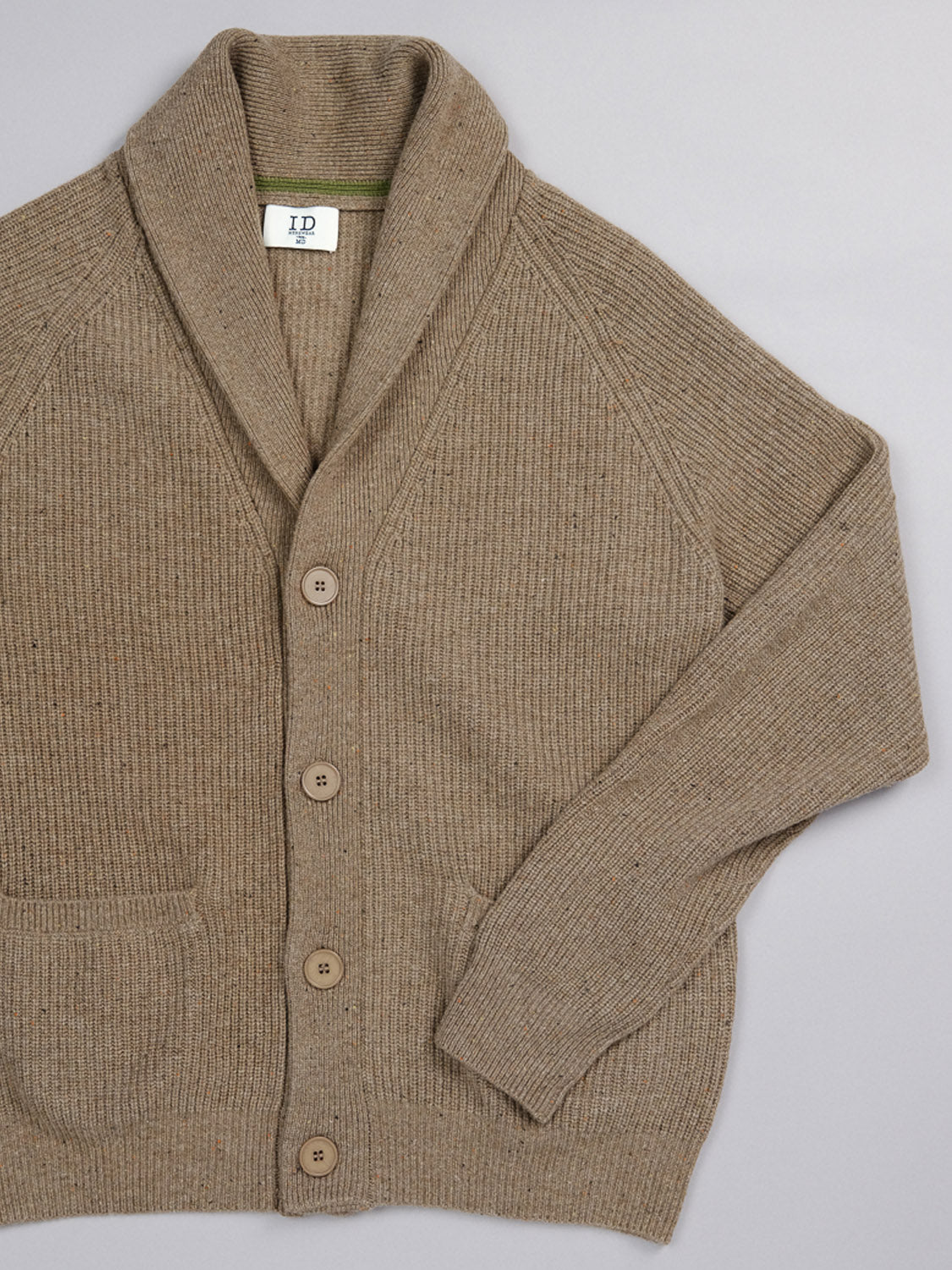 The Montreal Merino Wool Cardigan by ID Menswear is a beige piece crafted from speckled yarn with long sleeves, a shawl collar, and button-up front. Laid flat, it reveals two front pockets and ribbed texture details that highlight its fine merino wool composition.