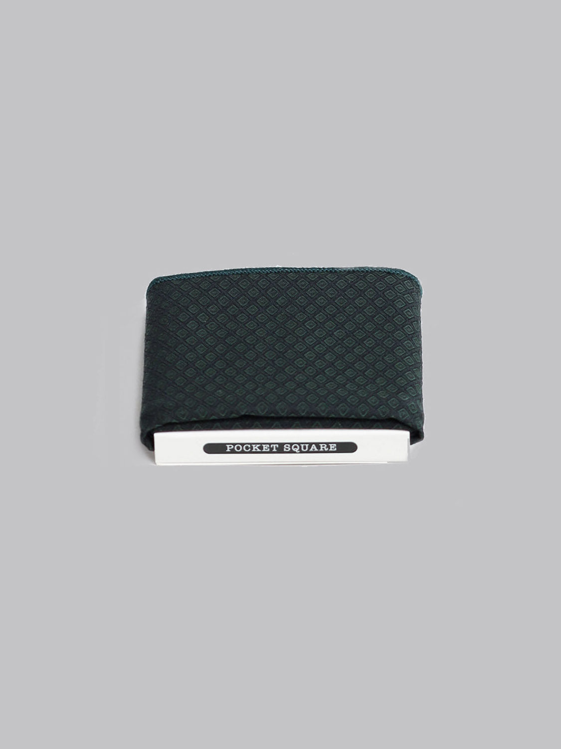 The Moroc Pocket Square by ID Menswear, handmade in Brooklyn from 100% cotton with a dark green pattern, is neatly folded and partially tucked into white packaging labeled POCKET SQUARE on a plain gray background.