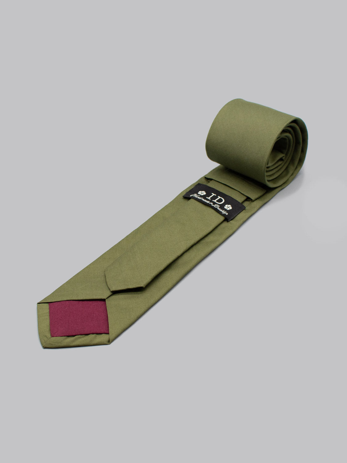 A rolled-up Liberty of London Tana Lawn™ Moss Tie by ID Menswear, handmade in Brooklyn, has an olive green design with a maroon tip and lies on a light gray background. The black lining label displays white text and symbols.