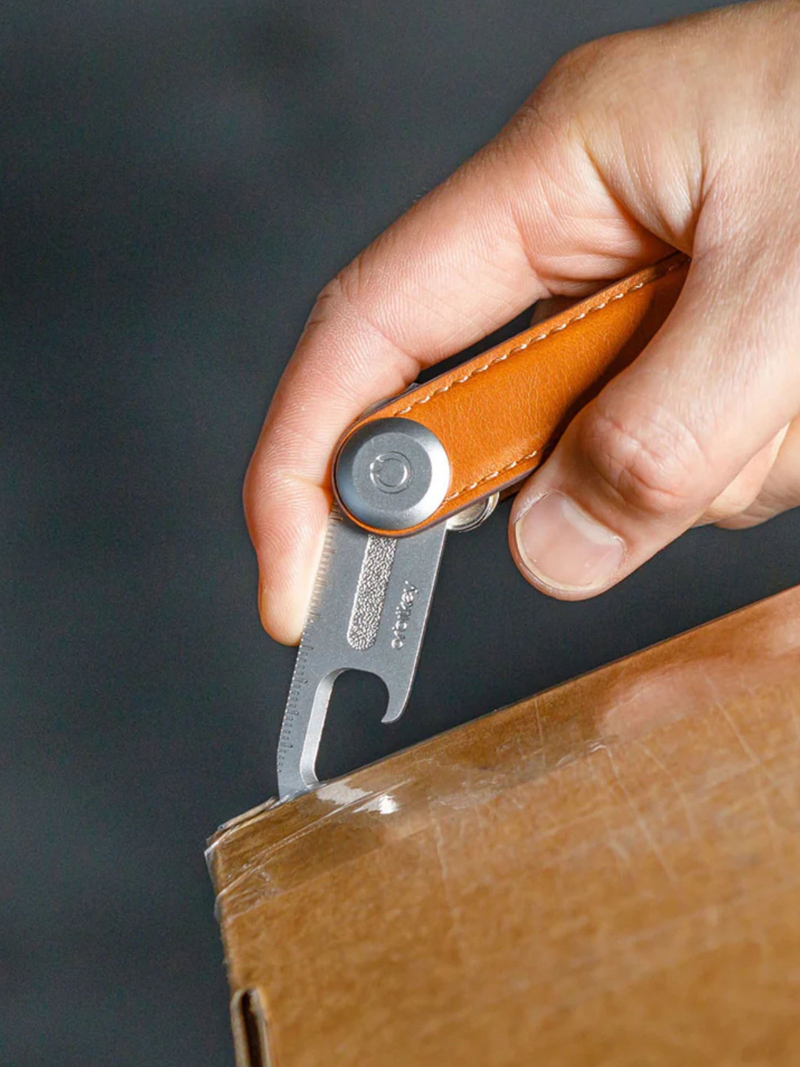 A hand uses the Orbitkeys Multi Tool V2 – A Handy Little Tool, featuring a stainless steel blade and leather handle, to cut open a cardboard box by sliding the blade under the tape at the box’s edge.