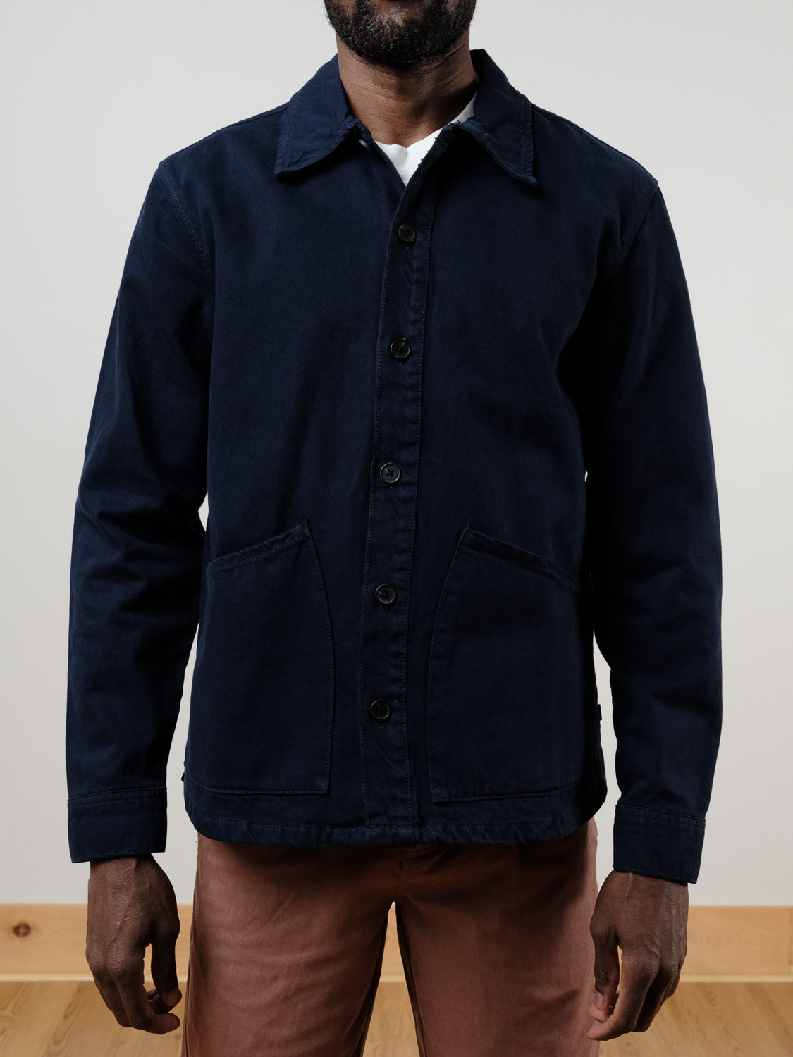 A man stands against a plain light background wearing the !SOLID Nadil Cotton Button-Up Overshirt in dark blue over a white shirt and brown pants. The overshirt features a collar and two large front pockets.