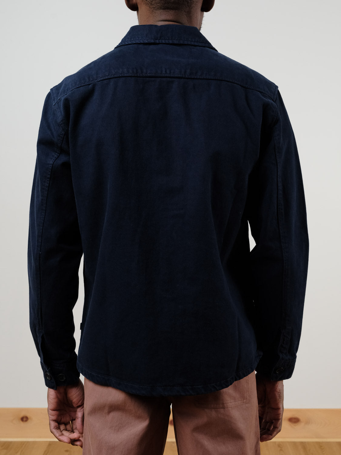 A person faces away from the camera, wearing the !SOLID Nadil Cotton Button-Up Overshirt in dark blue and light brown pants, set against a plain background with a wooden floor.